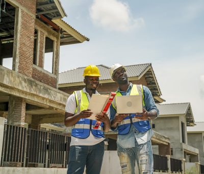 Two construction workers in safety gear analyze blueprints at a construction site. They stand in front of unfinished buildings, discussing and planning the next steps in sunny weather.